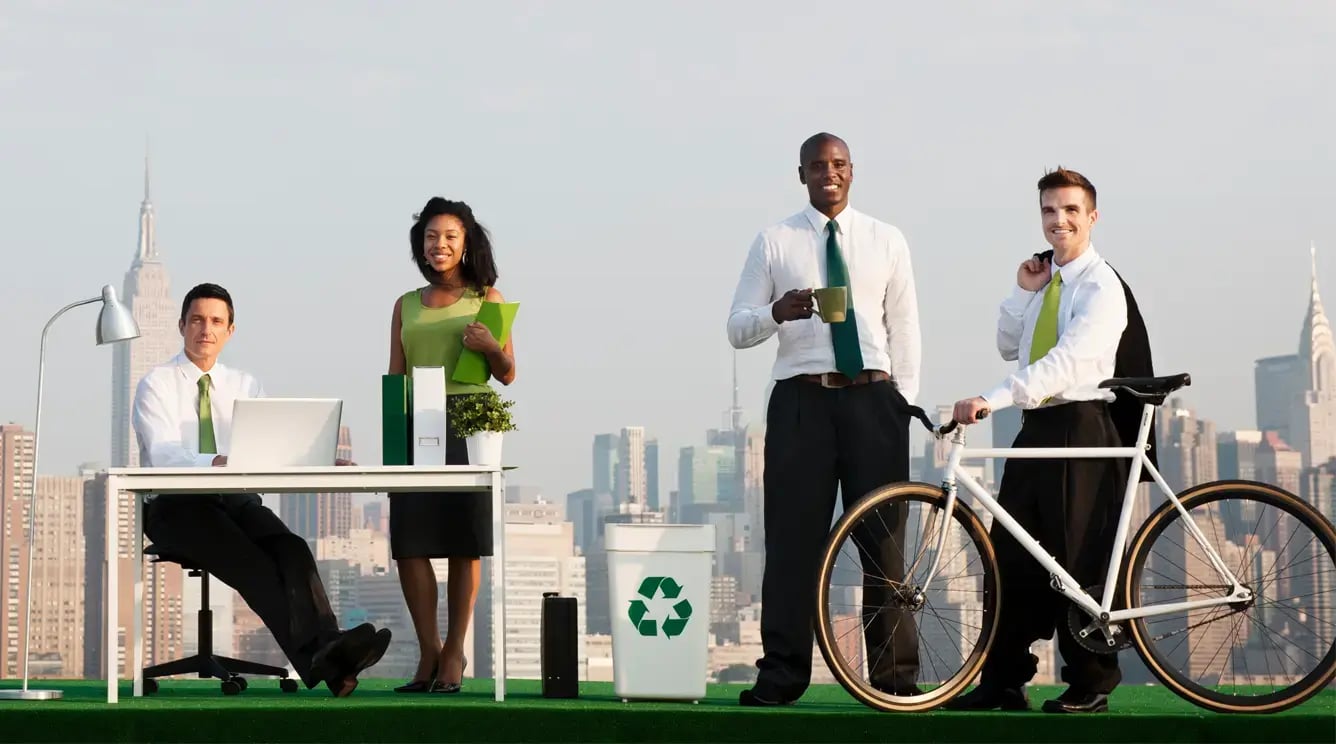 Office workers with green accessories pose in front of a city skyline with a desk, recycling bin and a bike. Office workers with green accessories pose in front of a city skyline with a desk, recycling bin and a bike.