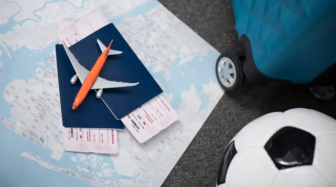 A map laid out on the floor with two passport and plane tickets on top. A small toy aeroplane balances on top of the passports. To the side, the corner of a wheeled suitcase and a football.